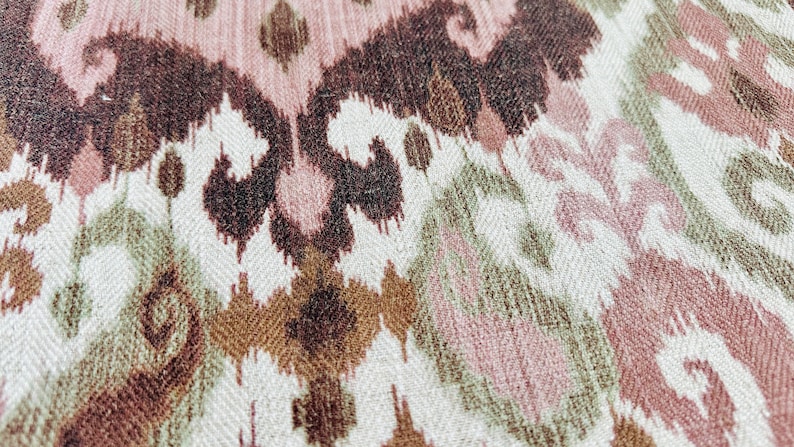May include: Close-up of a woven fabric with an ikat pattern. The design features repeating shapes in shades of brown, pink, and green on a cream background. The fabric has a textured appearance.