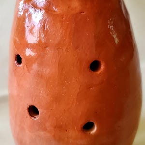 Può includere: Un ocarina in terracotta con sei fori, forma rotonda e una finitura liscia leggermente lucida.