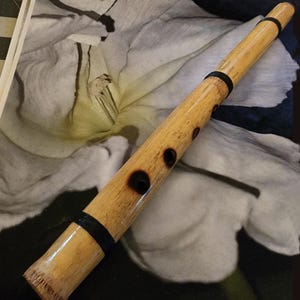 Puede incluir: Una flauta de bambú de color marrón claro con un cuerpo cilíndrico liso y varios orificios para los dedos. El instrumento está acentuado con tres bandas negras distintivas envueltas alrededor de la madera a intervalos a lo largo.