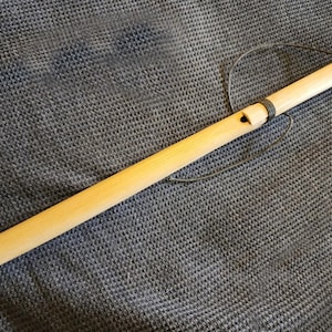 Puede incluir: Una flauta de madera cilíndrica de color claro con un acabado liso, que presenta un solo orificio de embocadura cerca de la parte superior y un cordón oscuro envuelto alrededor del cuerpo. Un hilo negro fino está unido a la flauta.