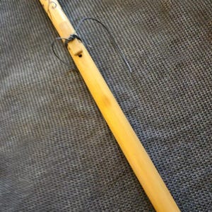 Puede incluir: Una flauta de madera de color claro con una cuerda oscura enrollada alrededor. La flauta tiene un pequeño agujero y algunas marcas cerca de la parte superior. El fondo es una tela gris texturizada.