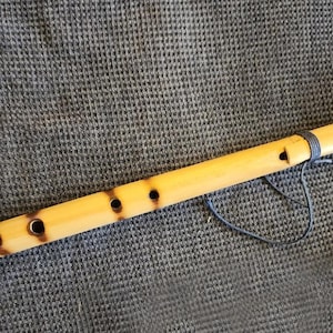 Puede incluir: Una flauta de bambú marrón claro con varios agujeros para los dedos y una banda oscura cerca de la parte superior. El instrumento tiene un acabado liso y pulido y un cordón oscuro adjunto. La flauta está sobre una superficie gris texturizada.
