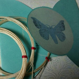 Op de afbeelding: Een spoel wit garen met rode accenten, gewikkeld rond een turquoise achtergrond. Een ronde kaart met een blauw vlinderontwerp is bevestigd aan de achtergrond.