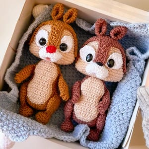 Può includere: Due bambole di scoiattolo a uncinetto fatte a mano in una scatola bianca con una coperta blu all'uncinetto. Uno scoiattolo è marrone e marrone chiaro con un naso rosso, l'altro è marrone e beige. La scatola è tenuta da una persona che indossa un maglione grigio.