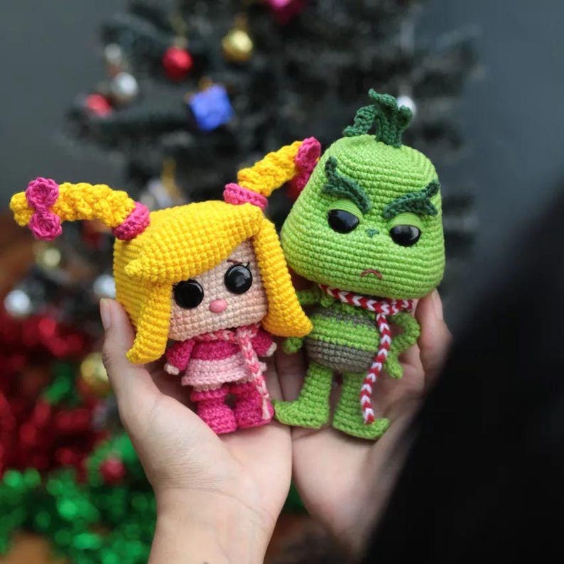 May include: Two crocheted dolls: a yellow-haired girl with pink accents and a green Grinch character. The girl doll has a pink scarf and dress, while the Grinch wears a red and white scarf. Both dolls have black eyes and are handmade.