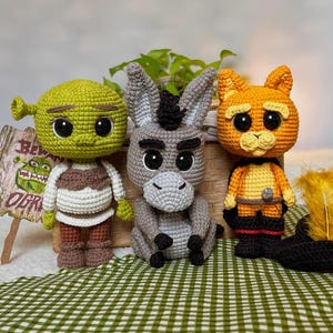 Peut inclure: Trois poupées au crochet faites à la main : Shrek, l'Âne et le Chat Potté. Shrek est vert avec une tenue blanche et marron. L'Âne est gris. Le Chat Potté est orange et noir. Elles sont disposées sur une surface à carreaux verts et blancs.