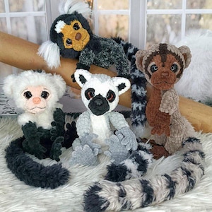 May include: A group of handmade monkey plush toys in different colours and patterns. The monkeys include a white and black monkey, a grey and white lemur, a brown monkey, and a black and white monkey. They are arranged on a white, fluffy surface.