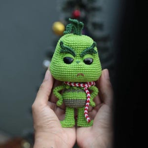 May include: A crocheted green character with a grumpy expression, black eyes, and a red and white scarf. The figure has a bulbous head, small arms, and legs. The character is a popular holiday figure.