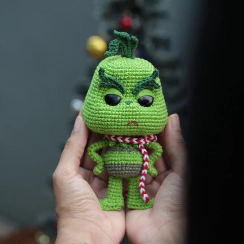May include: A crocheted green character with a grumpy expression, black eyes, and a red and white scarf. The figure has a bulbous head, small arms, and legs. The character is a popular holiday figure.