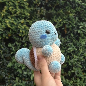 May include: A light blue crocheted plush toy with a round head, small arms and legs, and a brown and tan shell. The toy has a blue cheek and black eyes. The plush is held in a hand against a green leafy background.