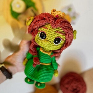 May include: Crocheted doll featuring a green face and body, red hair, and a gold crown. The doll wears a green dress with a yellow belt and black eyes. The doll is handmade and appears to be a character from a popular animated movie.