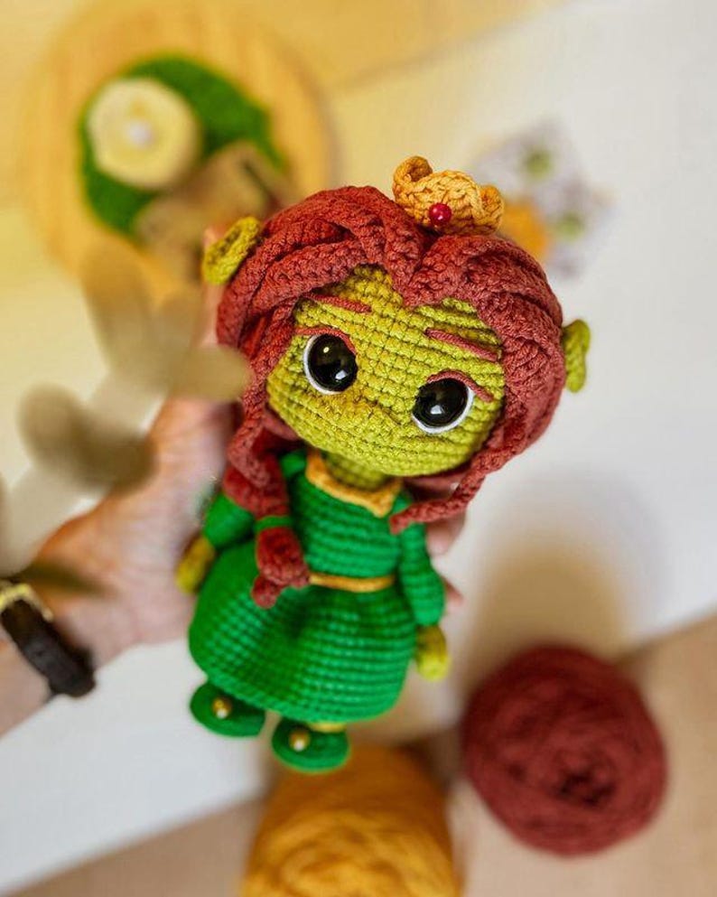 May include: Crocheted doll featuring a green face and body, red hair, and a gold crown. The doll wears a green dress with a yellow belt and black eyes. The doll is handmade and appears to be a character from a popular animated movie.