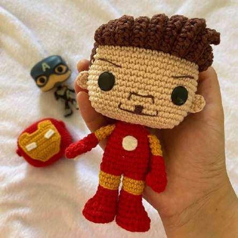 May include: Crocheted Iron Man doll in red, yellow, and beige. The doll has a square head with black eyes and brown hair. A Captain America figurine and Iron Man mask are also visible.