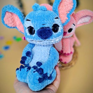 May include: A handmade blue plush toy with large ears and dark blue accents. The plush toy has large black eyes and is sitting in front of a pink plush toy. The plush toys are made of a soft, textured material.