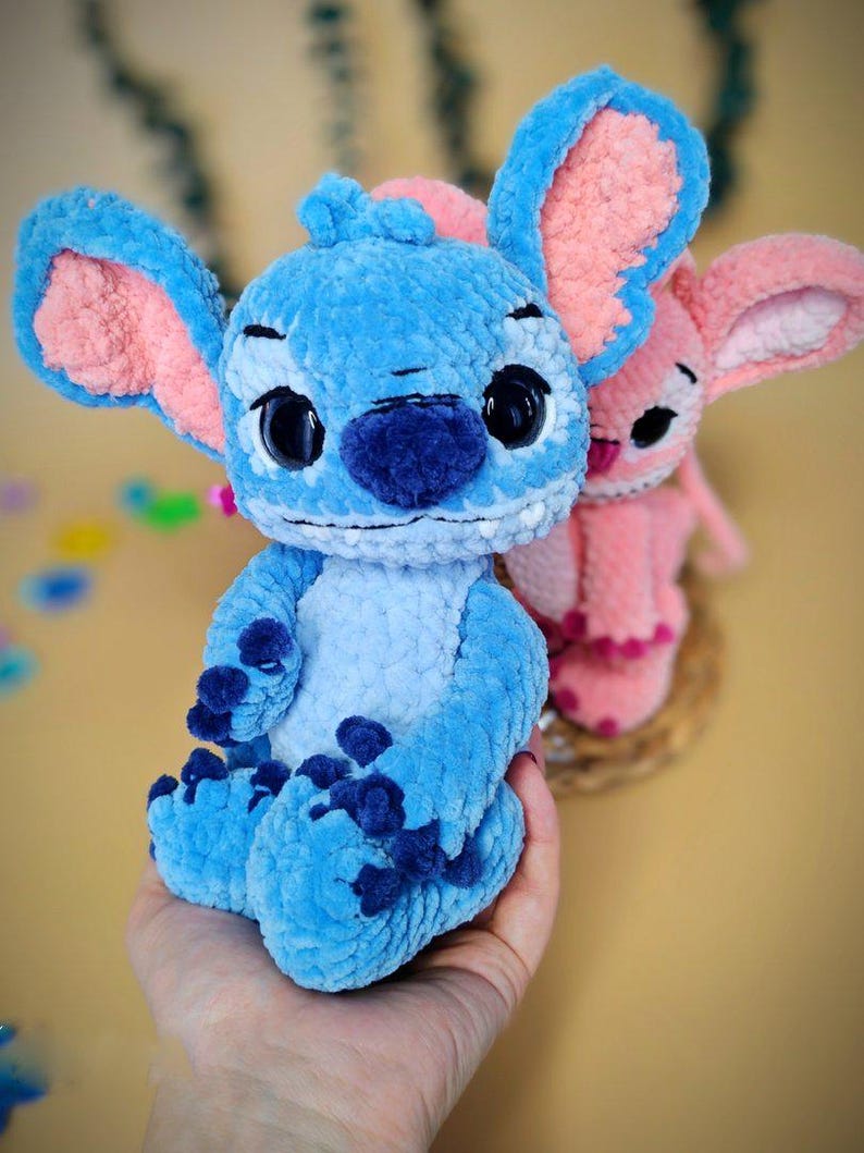 May include: A handmade blue plush toy with large ears and dark blue accents. The plush toy has large black eyes and is sitting in front of a pink plush toy. The plush toys are made of a soft, textured material.