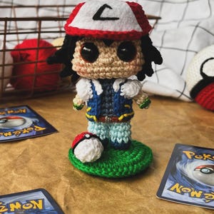 May include: Crocheted figure of a character wearing a red and white hat with a black "L" on it, a blue vest, and light blue pants. A crocheted Pokeball sits on a green base. Pokemon trading cards are in the background.