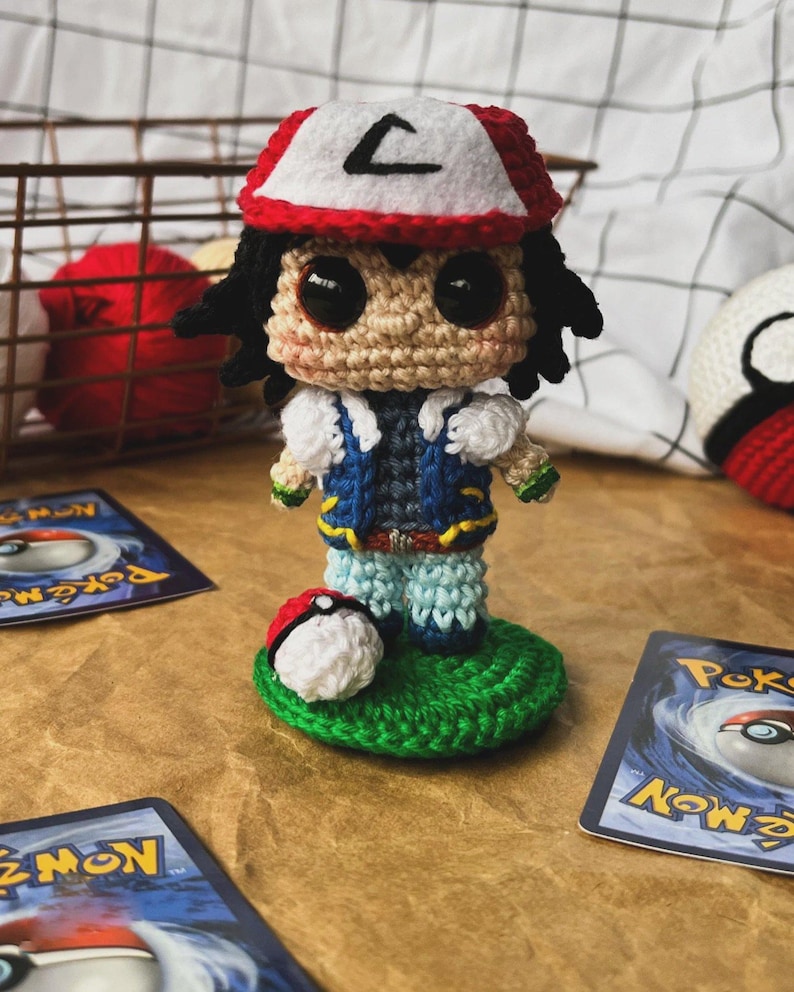 May include: Crocheted figure of a character wearing a red and white hat with a black "L" on it, a blue vest, and light blue pants. A crocheted Pokeball sits on a green base. Pokemon trading cards are in the background.