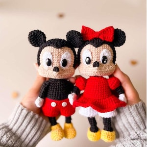 Può includere: Due bambole all'uncinetto fatte a mano, una maschile e una femminile, che assomigliano a personaggi dei cartoni animati. La bambola maschile indossa pantaloncini neri, una camicia nera e scarpe gialle. La bambola femminile indossa un vestito rosso e un fiocco.