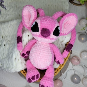 May include: A handmade pink crochet plush toy, resembling a cartoon character, with large ears, black eyes, and purple accents. The plush is seated in a woven basket, with a soft white blanket in the background. The toy is a light pink color.