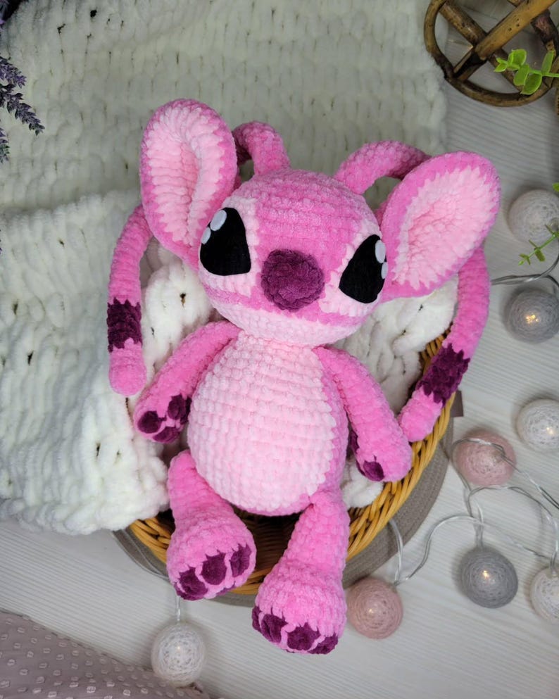 May include: A handmade pink crochet plush toy, resembling a cartoon character, with large ears, black eyes, and purple accents. The plush is seated in a woven basket, with a soft white blanket in the background. The toy is a light pink color.