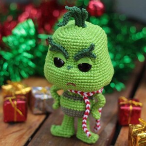 May include: A green crocheted Grinch doll with black eyes, a red and white scarf, and a grumpy expression. The doll is standing on a wooden surface with wrapped gifts in the background. The Grinch is a popular Christmas character.