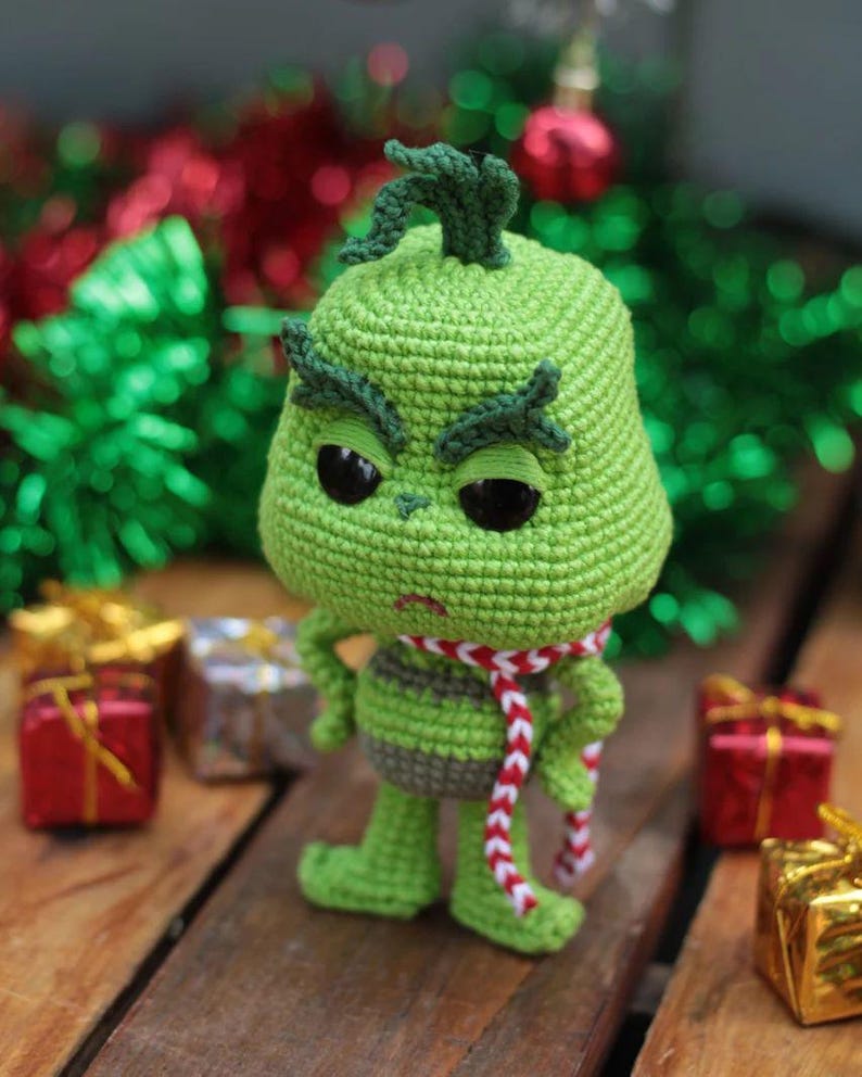 May include: A green crocheted Grinch doll with black eyes, a red and white scarf, and a grumpy expression. The doll is standing on a wooden surface with wrapped gifts in the background. The Grinch is a popular Christmas character.