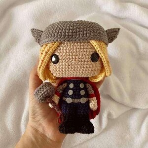 May include: A handmade crochet doll of Thor, featuring a gray helmet with wings, blonde yarn hair, and a red cape. The doll is holding a gray crochet hammer. The doll's outfit includes a navy blue vest with white circles.