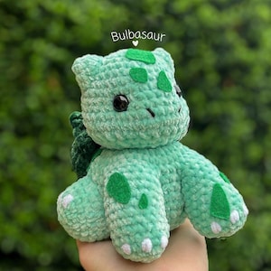 Puede incluir: Peluche de Bulbasaur de crochet verde con ojos negros y detalles blancos. El juguete está sentado en una mano.