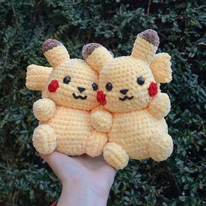 May include: Two crocheted yellow Pikachu plushies with brown ears and red cheeks. The plushies are sitting on a hand.