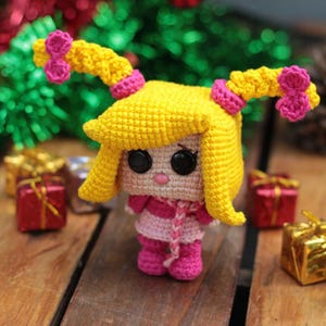 May include: A handmade crochet doll with yellow hair styled in two pigtails, each adorned with pink bows. The doll wears a pink and white striped dress, a pink scarf, and pink boots. Black button eyes and a small pink mouth complete the design.
