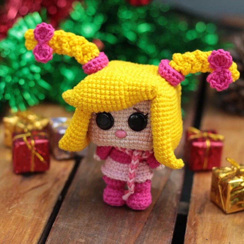 May include: A handmade crochet doll with yellow hair styled in two pigtails, each adorned with pink bows. The doll wears a pink and white striped dress, a pink scarf, and pink boots. Black button eyes and a small pink mouth complete the design.