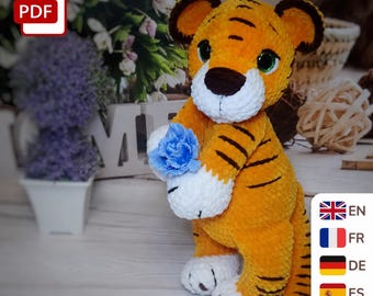 May include: A plush tiger toy made of orange and white yarn with dark brown stripes, large dark eyes, and rounded ears. The tiger is holding a small, textured blue flower in its front paws. The toy has white paws and a white muzzle area.