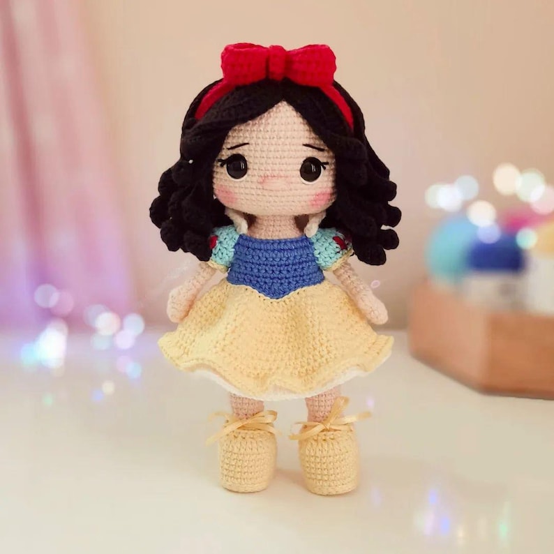 May include: A crocheted Snow White doll with black hair, a red bow, and a yellow dress with blue accents.