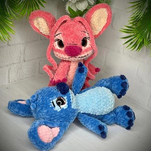 May include: Two plush toys, one pink and one blue, resembling cartoon characters. The pink toy has large ears and a prominent nose. The blue toy is lying down with a light blue belly and pink accents.