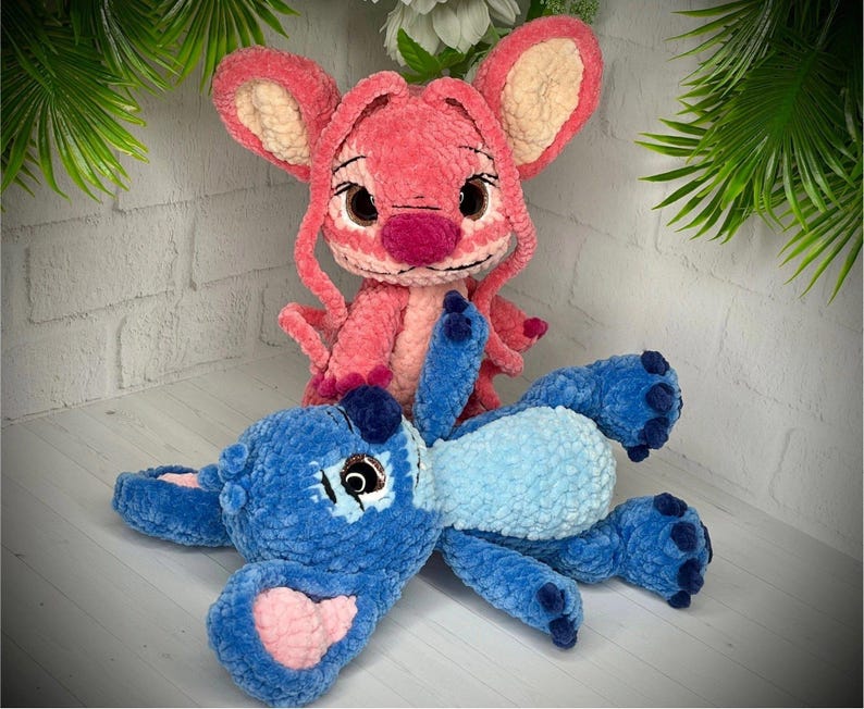 May include: Two plush toys, one pink and one blue, resembling cartoon characters. The pink toy has large ears and a prominent nose. The blue toy is lying down with a light blue belly and pink accents.