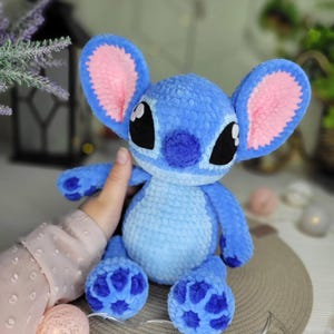 May include: A handmade blue plush toy with large ears, black eyes, and a small nose. The ears have pink inner linings. The toy is a character from a popular animated film. The plush toy is soft and cuddly.