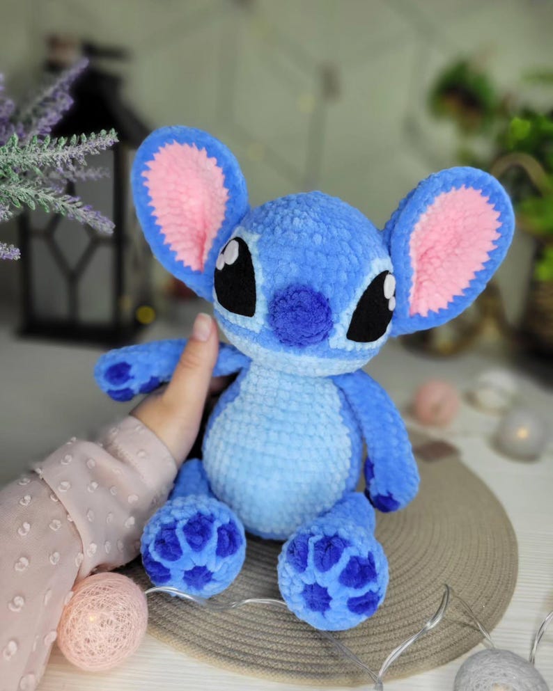May include: A handmade blue plush toy with large ears, black eyes, and a small nose. The ears have pink inner linings. The toy is a character from a popular animated film. The plush toy is soft and cuddly.