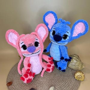 May include: Two plush toys, one pink and one blue, resembling a cartoon character. The pink toy has a purple nose and red accents on its feet. The blue toy has black eyes and a blue nose. Both toys have large ears with pink interiors.