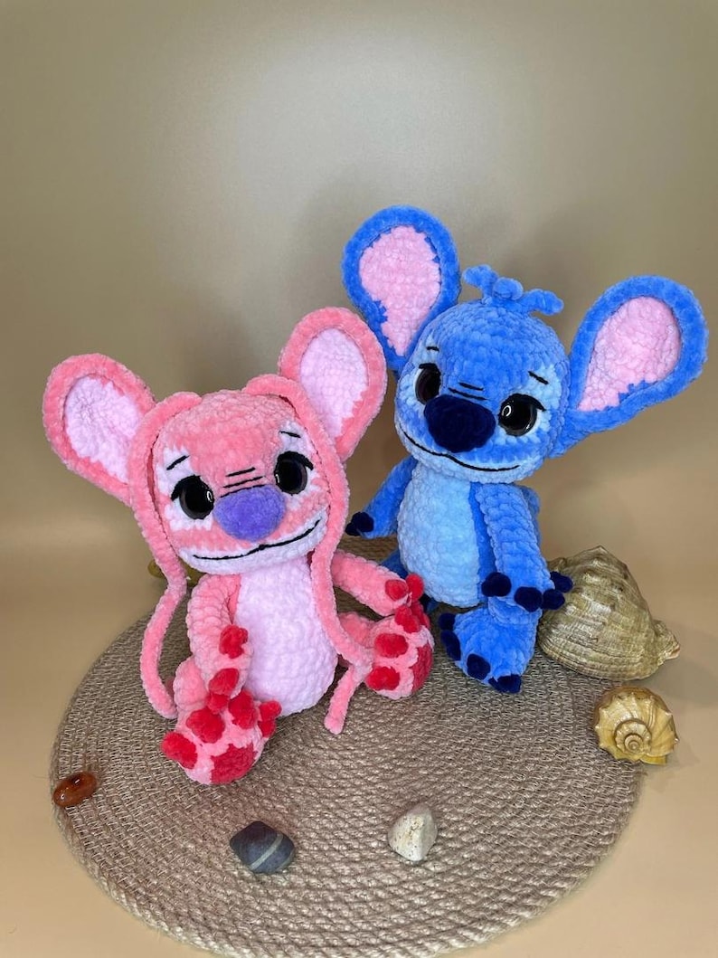 May include: Two plush toys, one pink and one blue, resembling a cartoon character. The pink toy has a purple nose and red accents on its feet. The blue toy has black eyes and a blue nose. Both toys have large ears with pink interiors.