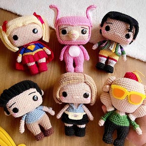 Puede incluir: Una colección de seis muñecas de ganchillo hechas a mano. Las muñecas presentan varios atuendos y peinados, incluyendo un disfraz de superhéroe, un traje de conejito rosa y otros diseños inspirados en personajes. Las muñecas están hechas con hilo y tienen ojos de botón negros.
