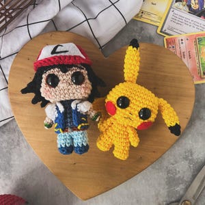 May include: Crocheted figures of Ash Ketchum and Pikachu are displayed on a wooden heart-shaped board. Ash wears a red and white hat, blue vest, and blue pants. Pikachu is yellow with red cheeks. Pok&eacute;mon cards and scissors are also visible.