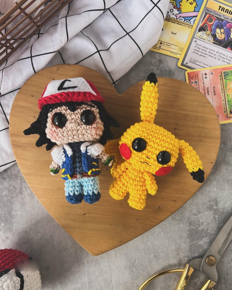 May include: Crocheted figures of Ash Ketchum and Pikachu are displayed on a wooden heart-shaped board. Ash wears a red and white hat, blue vest, and blue pants. Pikachu is yellow with red cheeks. Pok&eacute;mon cards and scissors are also visible.