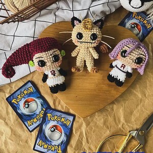 May include: Crocheted Pok&eacute;mon characters and a Pok&eacute; Ball sit on a wooden heart. The characters include a Meowth, and two Team Rocket members. Two Pok&eacute;mon trading cards and a pair of gold scissors are also visible.