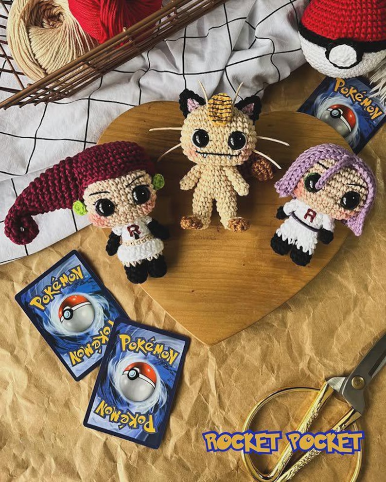 May include: Crocheted Pok&eacute;mon characters and a Pok&eacute; Ball sit on a wooden heart. The characters include a Meowth, and two Team Rocket members. Two Pok&eacute;mon trading cards and a pair of gold scissors are also visible.