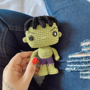 May include: A handmade green crochet doll of a superhero with black hair, black eyes, and purple shorts. The doll is held in a person's hand against a background of blue jeans.