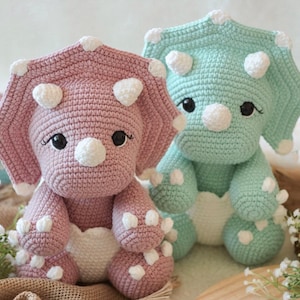 Peut inclure: Deux dinosaures triceratops au crochet faits à la main. L'un est rose, l'autre vert menthe. Les deux ont des accents blancs sur leurs volants, leurs cornes et leurs ventres. Ils ont des yeux noirs et sont assis droits.