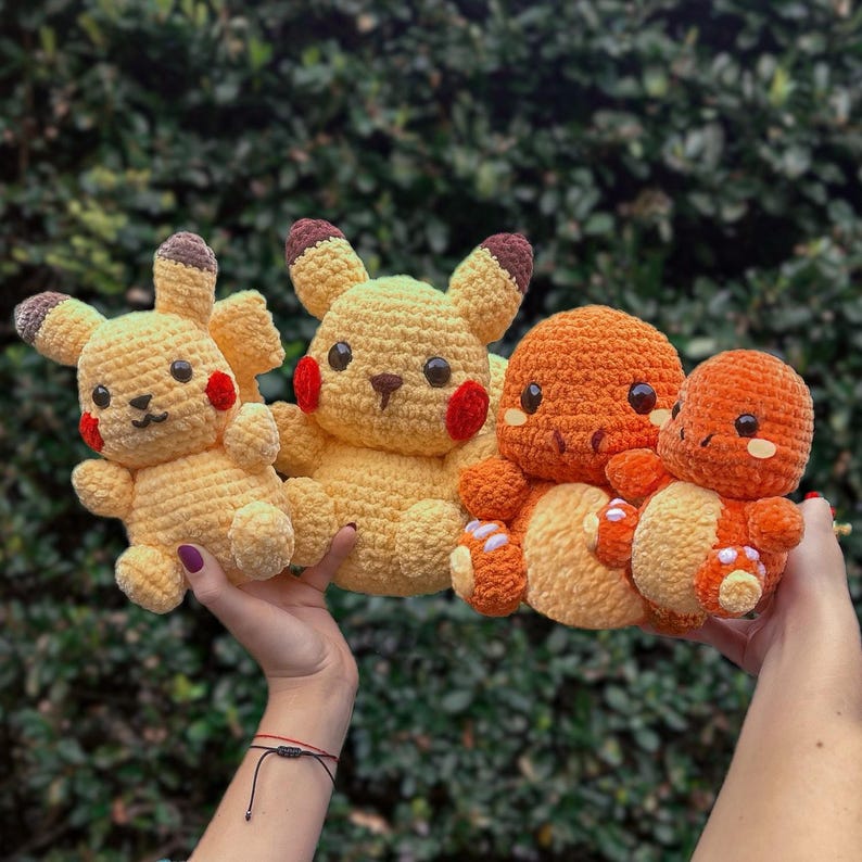 May include: Four crocheted plush toys, two yellow Pikachu and two orange Charmander, are held in the hands of a person. The toys are all smiling and have big eyes.