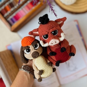 May include: Two handmade crochet toys: a brown dog wearing an orange hat and a red boar with a black tuft. The dog has a beige body and the boar has a red body with black accents. Both have large, expressive eyes.