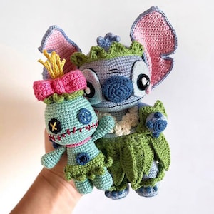 Peut inclure: Deux poupées crochetées à la main. L'une est un personnage bleu clair avec de grandes oreilles, un nez bleu et une jupe verte. L'autre est une poupée plus petite avec un nœud rose, une jupe verte et des traits du visage cousus. Les poupées sont tenues sur un fond blanc.