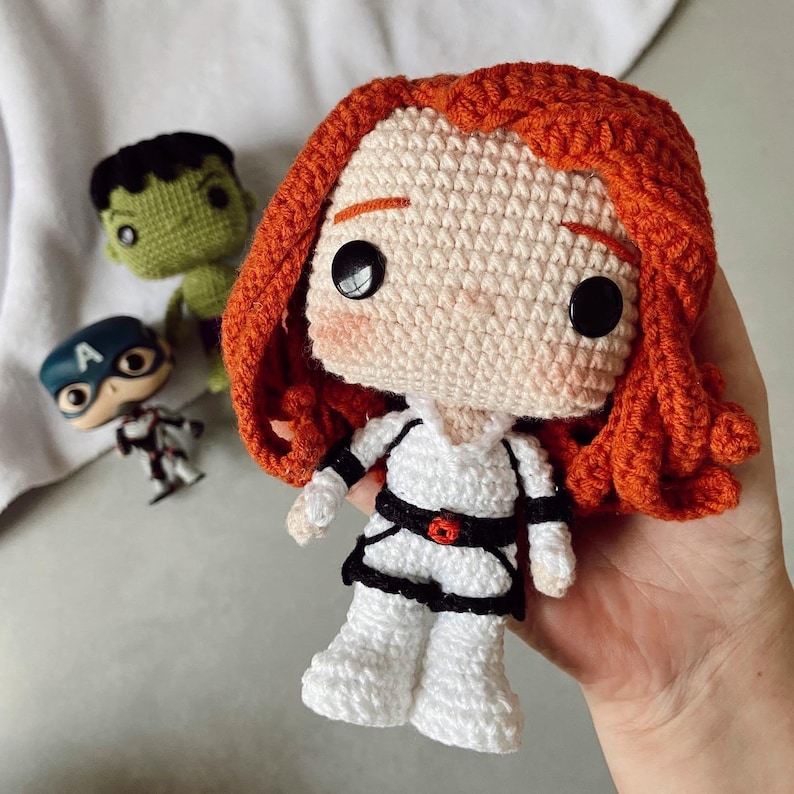 May include: Crocheted doll of a character with red hair, wearing a white outfit with black accents and a red belt. Other small character dolls are in the background, including a green Hulk and a Captain America.